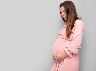 Pregnant woman in dress holds hands on belly on a grey background. Pregnancy, maternity, preparation and expectation concept. Close-up, copy space. Beautiful tender mood photo of pregnancy.