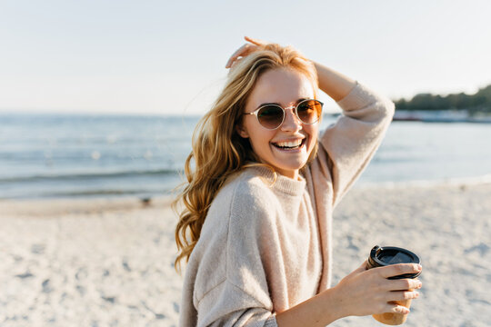 Winsome woman in sunglasses spending cold morning beside sea. Blithesome blonde lady with cup of coffee laughing at ocean coast