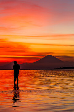 Man stands in the water silhouetted against a volcano at sunset