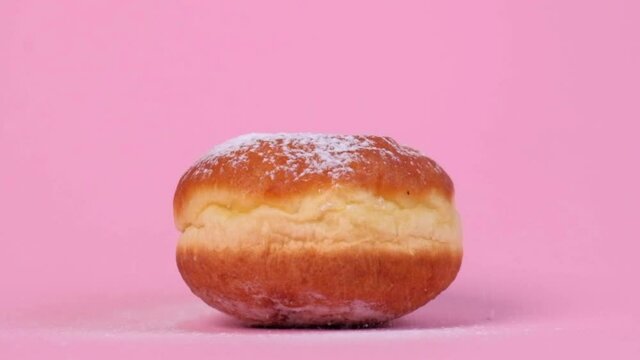 Nice round donut with filling. Powdered sugar flies from above. On a pink background. Pastry product, contains a lot of sugar.