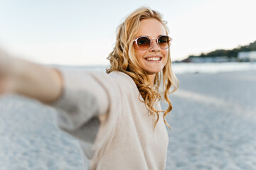 Cute woman with wavy blond hair sincerely smiles and takes selfie at sea