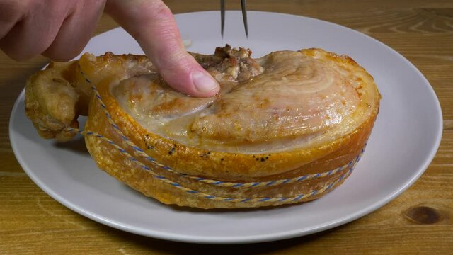 Closeup POV shot of a man’s finger sliding a joint of hot, juicy, joint of roast pork, with crisp crackling and holding string on the outside, from a carving fork onto a plate.