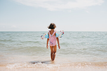 Toddler girl wearing pink swimsuit with unicorns and kids' swimming armbands on the sea
