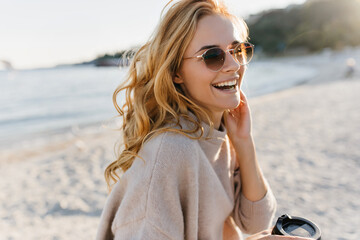 Cute woman genuinely laughs, relaxing on beach. Blonde girl in glasses and sweater holds cup of coffee