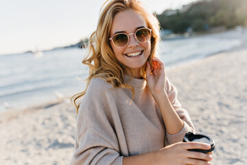 Gorgeous blonde woman in sunglasses and sweater standing at sea coast. Refined curly girl drinking...