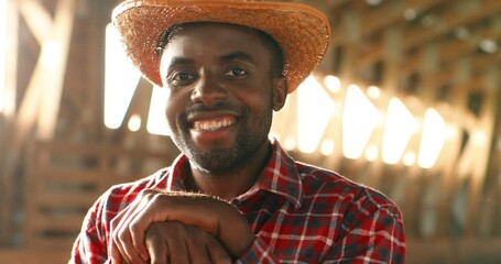 Portrait of young handsome African American man farmer in hat smiling at camera and leaning on pitchfork in barn. Good-looking cheerful male shepherd smile in dark stable. Close up. Flies flying. - Powered by Adobe