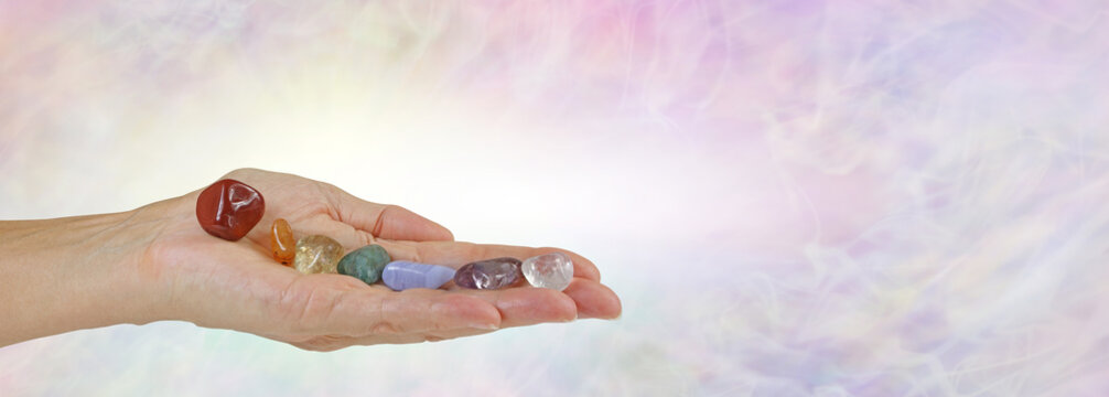 Seven Chakra Stones Message Banner - open flat female hand with seven tumbled stones neatly balanced in a row against a pale wispy energy formation background with copy space

