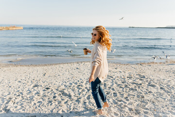 Charming lady in jeans and sweater looking at sea. Outdoor portrait of adorable girl holding cup of coffee during photoshoot near ocean