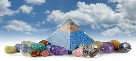 Orgone pyramid power and healing crystals - copper spiral and clear quartz on top of blue chip stones inside Orgone pyramid surrounded by mixed tumbled healing stones against blue sky background
