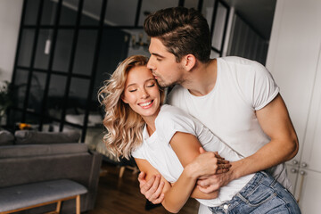 Romantic guy kissing wife. Indoor portrait of graceful curly girl embracing husband.