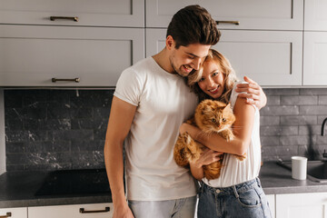 Pretty young woman holding cat during family photoshoot. Cute brunette man embracing his wife. © Look!
