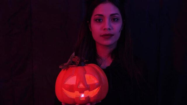 Close up, portrait of young pretty and sexy Asian woman in halloween concept costume, holding lantern, staring to camera, smile and beckoning over dark background with red and perple light setting
