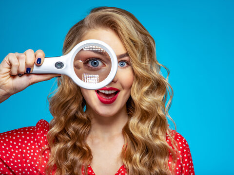 Surprised blonde holds a magnifying glass in front eye. Wonder young woman holds a magnifying glass in front of her eye. Funny girl with a bright smile and loupe. Eyesight care. Funny face of a girl