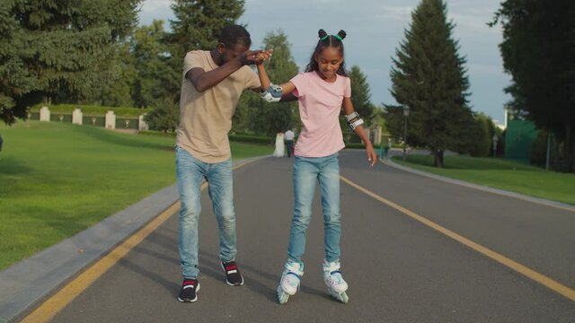 Caring affectionate african father securing happy mixed race teenage daughter, holding her hands helping to maintain balance on roller skates while teaching child rollerblading in summer park.