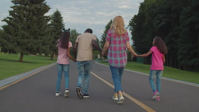 Rear view of carefree happy multiracial family with lovely elementary age daughters holding hands, walking away on park road, expressing unity, harmony, love and positivity during outdoor leisure.