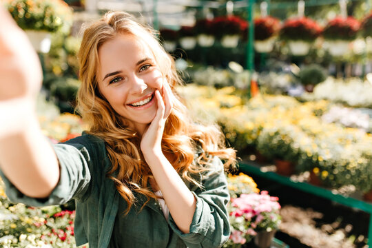 Sunny weather contributes to good photo of young woman with red hair. Girl with green eyes makes selfie against background of plants