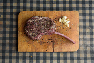 Dry aged raw Tomahawk beef steak piece place on wooden cutting board with herbs and ingredients...