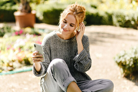 Girl In Gray Baggy Sweater Looks Into Her Phone And Selects Music To Listen To. Portrait Of Smiling Woman With Beautiful Curls