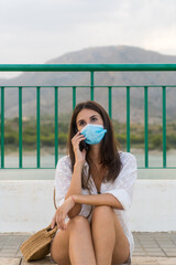 Girl with mask seated talking by phone at sunset in a mountain lake.