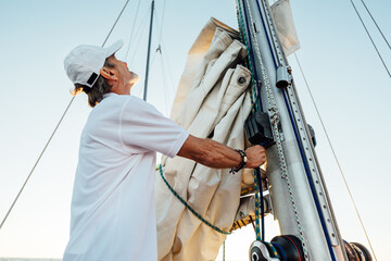 Mature captain looking up while adjusting sail. Senior yachtsman preparing a boat for a vacation...