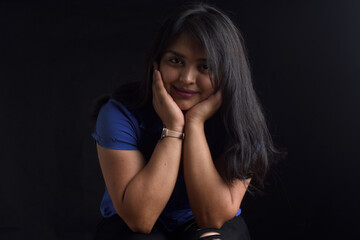portrait of a latin woman sitting on chair and looking at camera on black background, two hands on face