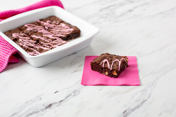 Pan of Homemade Brownies with Pink Frosting on a White Countertop with One Piece on a Pink Napkin in Front