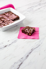 Pan of Homemade Brownies with Pink Frosting on a White Countertop with One Piece on a Pink Napkin in Front