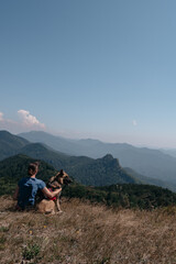 Fototapeta premium Dog traveler goes to the mountains with his best friend man and enjoys nature and beautiful scenery. A young traveller girl is sitting on top of a mountain with her German shepherd.