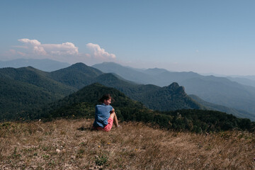 Naklejka premium Beautiful mountain and hilly landscape in warm, Sunny weather and the young tourist. A female traveler sits on top of a hill and looks at the surrounding mountains.