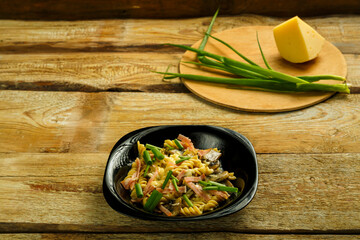 Black plate with pasta with ham and mushrooms in a creamy sauce on the table with a fork in the background.