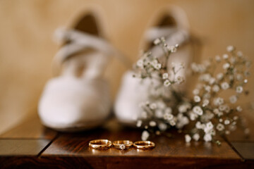 Wedding rings and an engagement ring on a wooden table with womens wedding white shoes and wildflowers.