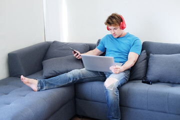 Young Caucasian man listening to musice by headphone and searching online social media with laptop. He is sitting on sofa in living room. Lifestyle and techonology on holiday Concept.