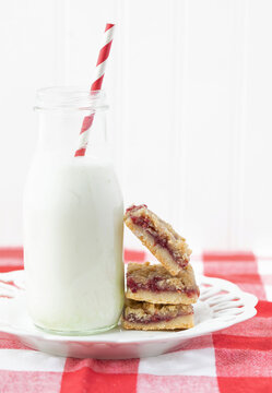 Original food photograph of homemade red raspberry streusel bars stacked on a white plate with a bottle of milk with a red and white straw sitting on a red and white checked napkin 