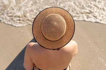 A young Caucasian girl is sitting on the sea sand on the beach in a straw hat. Rest. Selective focus. Rear view