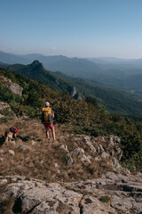 A dog traveler and its human owner with a backpack walk in the national Park and enjoy nature. A girl traveler and her German shepherd stand on a hill and look at the mountains around.