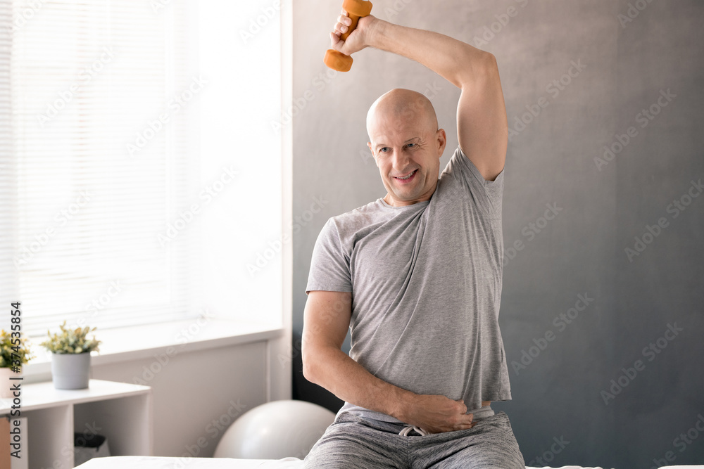 Poster bald middle aged man in activewear doing physical exercise with dumbbell