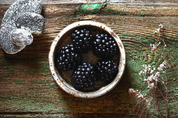 Fresh blackberries lie in a saucepan, next to dried flowers and textured stones, on a wooden background. The view from the top..