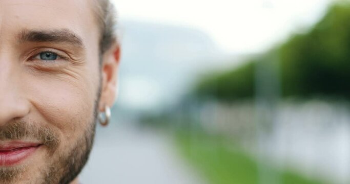 Close up of half face of young caucasian cheerful handsome man with earing in ear smiling to camera and looking straight outdoor. Portrait of happy stylish good-looking male.