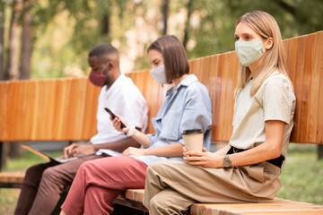Young blond bored woman in protective mask having coffee while sitting on bench