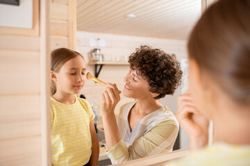 Pretty young smiling woman touching face of her cute little daughter with brush