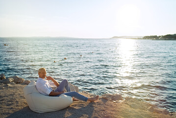Senior businessman sitting on deck chair on the beach during sunset