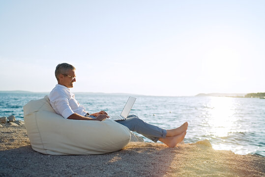 Senior man working on his laptop lying on deck chair on the beach during sunset