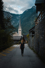 Fototapeta na wymiar A travel girl exploring the old town of Hallstatt on a rainy summer day. Street view with the famous old and historic chruch in the background. Austrian Alps, Salzkammergut in Austria, Europe