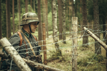 A soldier in an iron helmet of world war II, standing near a barbed wire in the woods. Military historical reconstruction.