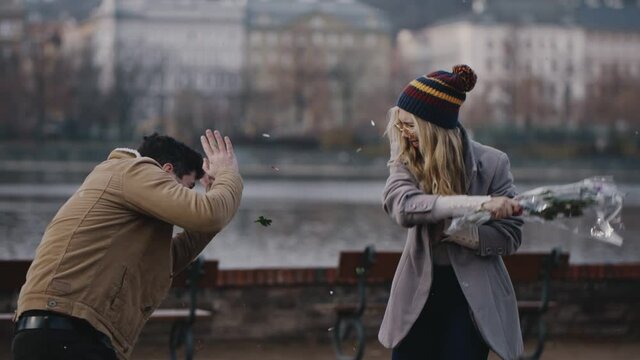Crazy caucasian girl taking bouquet of flowers and brutally hiting guy on river embankment. Young couple screaming and quarreling on date. Jealous woman beating scared man with bouquet after betrayal.