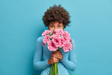 Good looking delicate African American woman smells rosy gerbera flowers, enjoys pleasant odour...