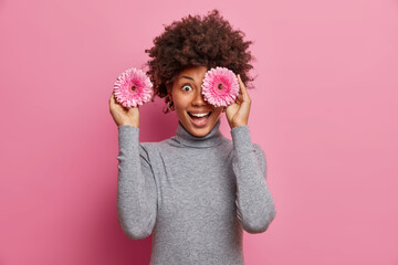 Horizontal shot of happy Afro American woman covers eyes with rosy gerbera flowers, has fun and...