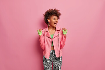 Overjoyed slim woman has Afro hair cleches fists with joy, feels very happy and lucky after training in gym, motivates to sport wears stylish pink jacket and sport gloves poses indoor exclaims loudly