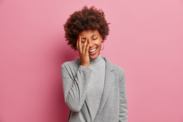 Fototapeta na wymiar Overjoyed dark skinned female entrepreneur laughs positively, stands with closed eyes, laughs at funny joke, makes face palm, dressed elegantly, shows white teeth, isolated over rosy background