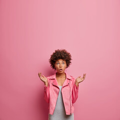 Vertical shot of stunned curly haired woman looks with great surprisement above, spreads palms and hears something unbelievable, wears pink jacket, blank copy space above for your information